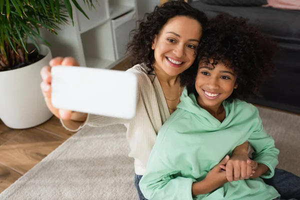 Vista de ángulo alto de la madre afroamericana feliz tomando selfie con alegre hija preadolescente en el teléfono inteligente - foto de stock