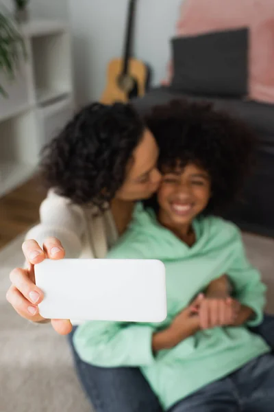 Vista de ángulo alto de la madre afroamericana rizada besar mejilla de la hija y tomar selfie en el teléfono inteligente enfocado - foto de stock