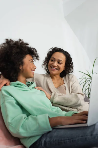 Alegre africano americano preadolescente chica usando laptop cerca feliz madre - foto de stock