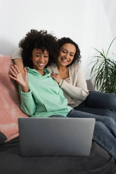 Feliz africano americano madre abrazando alegre preadolescente hija saludando mano durante la videollamada - foto de stock