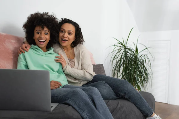 Feliz africano americano madre abrazando emocionado preadolescente hija viendo película en portátil - foto de stock