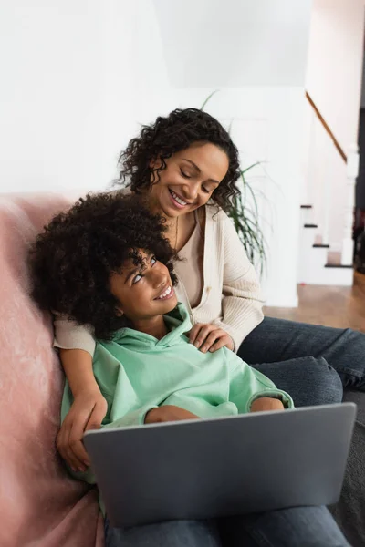 Happy african american mother hugging cheerful preteen daughter near laptop — Stock Photo