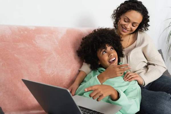 Happy african american mother looking at excited daughter lying with laptop on couch — Stock Photo