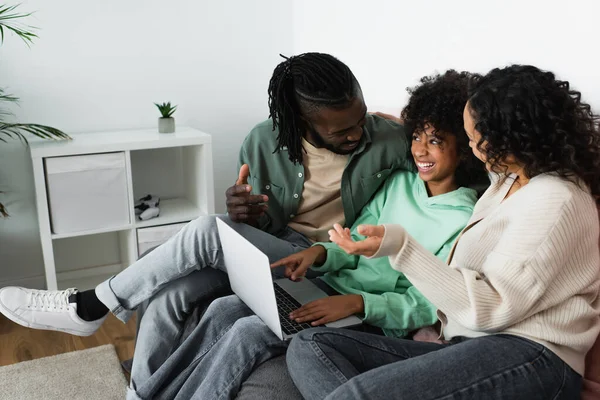 Feliz afroamericano padres mirando sonriente preadolescente hija con portátil sentado en sofá - foto de stock