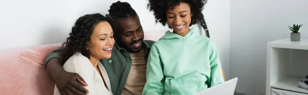 Щаслива афроамериканська сім'я дивиться комедійний фільм на ноутбуці у вітальні, банер — стокове фото