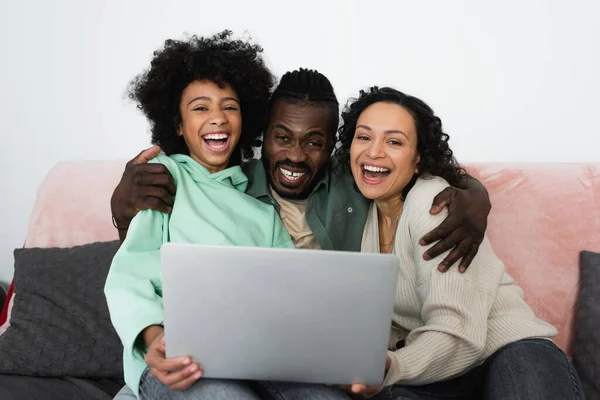 Familia afroamericana feliz mirando la cámara cerca de la computadora portátil en la sala de estar - foto de stock