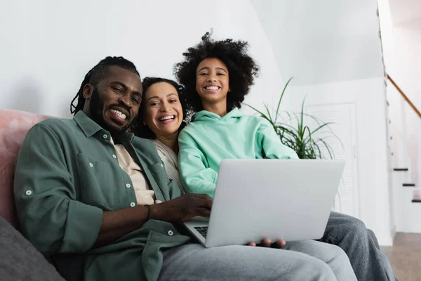 Joyeuse famille afro-américaine regardant la caméra près de l'ordinateur portable dans le salon — Photo de stock