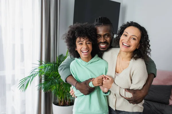 Feliz africano americano hombre abrazando alegre preadolescente hija y esposa en casa - foto de stock