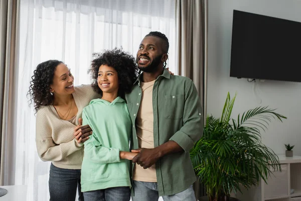 Smiling african american parents hugging cheerful preteen daughter at home — Stock Photo