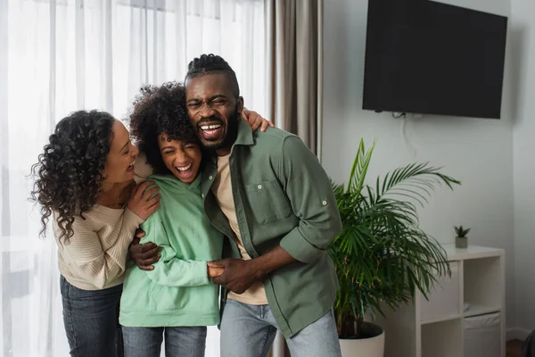 Feliz afroamericano padres riendo y abrazando alegre preadolescente hija en casa - foto de stock