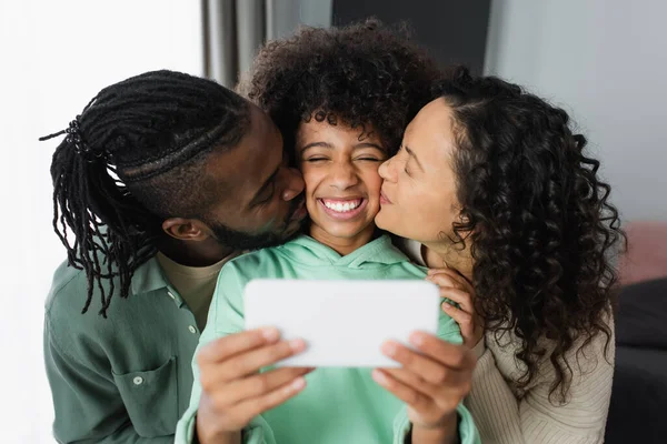 Glückliche afrikanisch-amerikanische Eltern küssen Wange ihrer Tochter, während sie Selfie zu Hause machen — Stockfoto