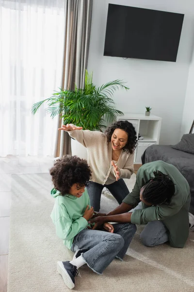 Fröhliche afrikanisch-amerikanische Vater kitzelt preteen Tochter in der Nähe glückliche Frau im Wohnzimmer — Stockfoto
