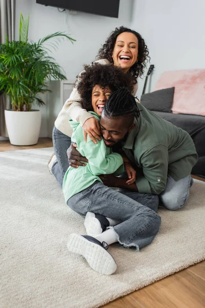 Веселый африканский американский отец щекочет дочь-подростка рядом с положительной женой в гостиной — стоковое фото