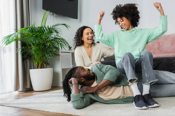 Positivo afroamericano preadolescente chica sentado en la cadera de padre y mostrando el gesto de poder cerca de feliz madre - foto de stock