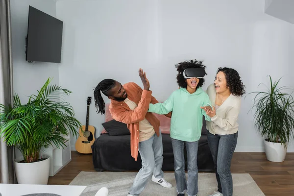 Padres afroamericanos positivos gestos cerca de preadolescente hija juego en auriculares vr en la sala de estar - foto de stock