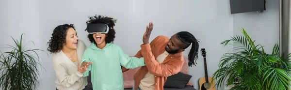 Padres afroamericanos positivos cerca del juego preadolescente de la hija en auriculares vr, bandera - foto de stock