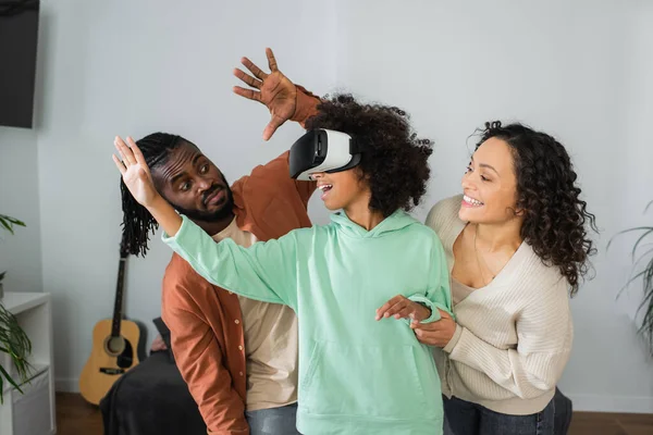 Africanos americanos padres sonriendo cerca preadolescente hija juego en vr auriculares en sala de estar - foto de stock
