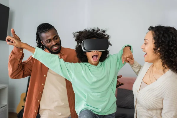 Padres afroamericanos felices tomados de la mano de la hija preadolescente sorprendida jugando en auriculares vr en la sala de estar - foto de stock