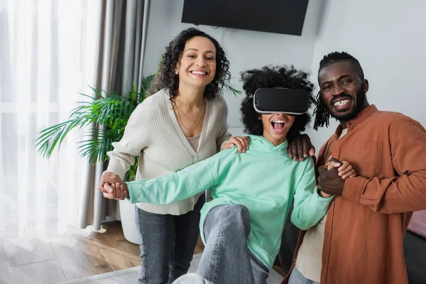 Alegres pais afro-americanos segurando as mãos de jogos de filha pré-adolescente espantados no fone de ouvido vr na sala de estar — Fotografia de Stock
