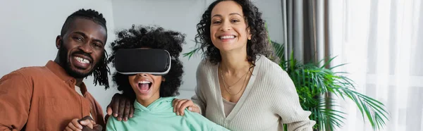 Cheerful african american parents near amazed preteen daughter gaming in vr headset, banner — Stock Photo