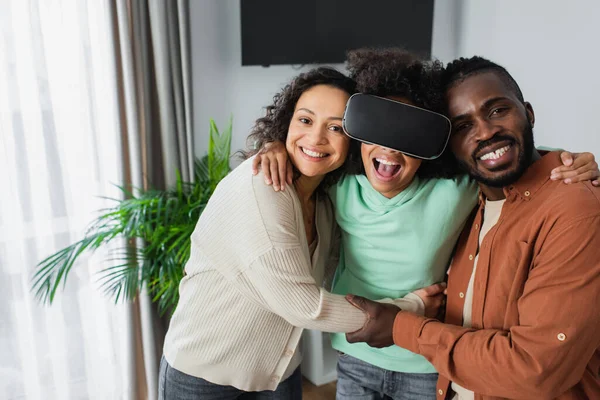 Feliz africano americano padres abrazando emocionado preadolescente hija juego en vr auriculares - foto de stock