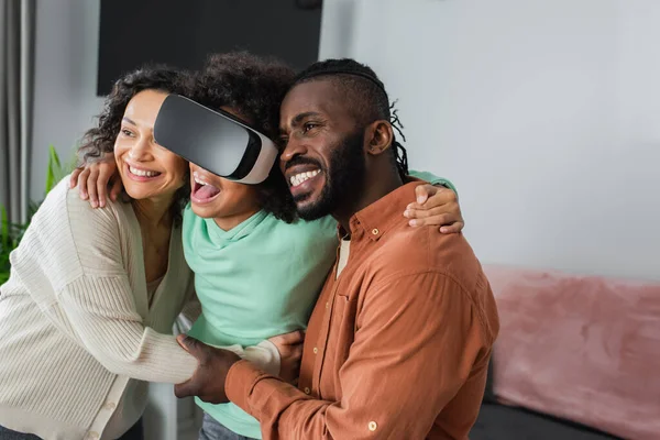 Alegre afroamericano padres abrazando emocionado preadolescente hija juego en vr auriculares - foto de stock