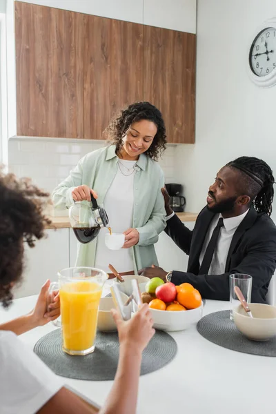 Feliz africano americano mujer verter café en taza cerca marido en traje y preadolescente hija durante el desayuno - foto de stock