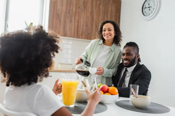 Feliz afroamericana mujer sosteniendo cafetera y taza cerca de marido en traje y preadolescente hija durante el desayuno - foto de stock