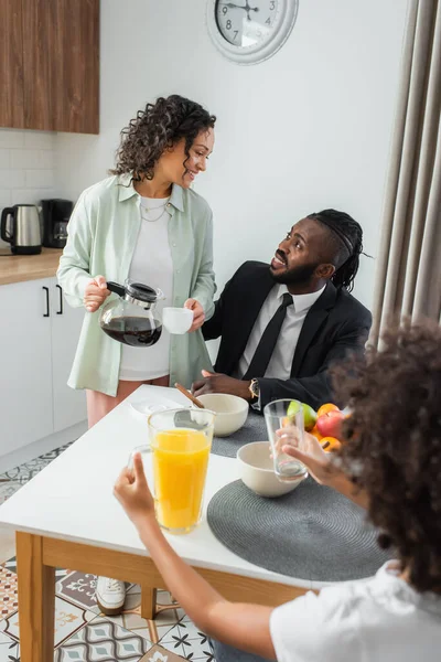 Feliz mulher afro-americana segurando panela de café e xícara enquanto olha para o marido de terno perto da filha pré-adolescente durante o café da manhã — Fotografia de Stock