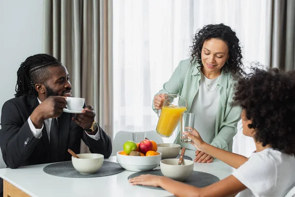 Glücklich afrikanisch-amerikanische Frau gießt Orangensaft im Glas in der Nähe Ehemann im Anzug und Frühchen Tochter während des Frühstücks — Stockfoto