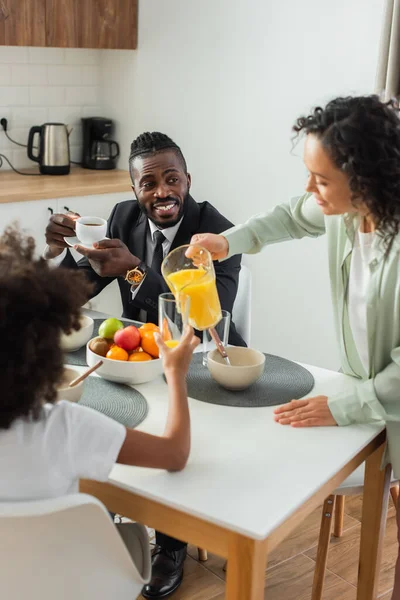 Afrikanisch-amerikanische Frau gießt Orangensaft in Glas neben glücklichem Ehemann im Anzug und frühgeborener Tochter während des Frühstücks — Stockfoto