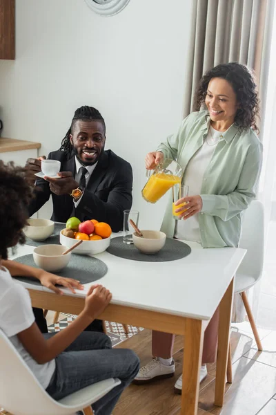 Cheerful african american woman pouring orange juice in glass near happy husband in suit and daughter during breakfast — Stock Photo