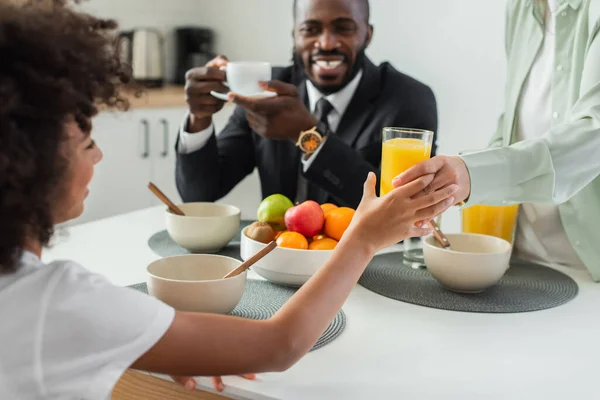 Африканская американская женщина дает стакан апельсинового сока счастливой дочери рядом с мужем во время завтрака — стоковое фото