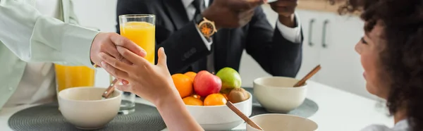 Africano americano mujer dando vaso de jugo de naranja a feliz hija cerca marido durante el desayuno, bandera - foto de stock