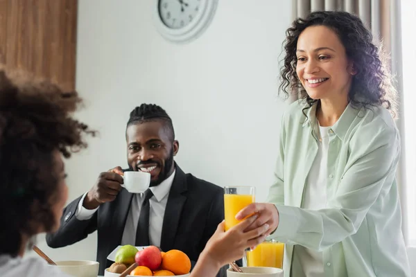 Fröhliche afrikanisch-amerikanische Frau gibt ein Glas Orangensaft an ihre glückliche Tochter in der Nähe ihres Mannes, der beim Frühstück Kaffee trinkt — Stockfoto