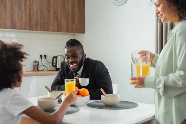 Cheerful african american businessman in suit laughing and looking at daughter near wife with jug of orange juice — Stock Photo