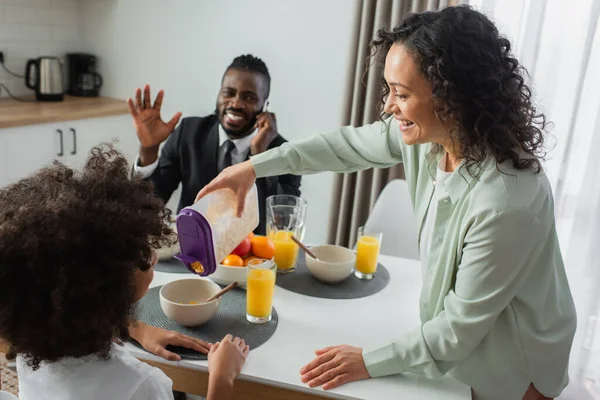 Allegra donna africana americana versando fiocchi di mais in ciotola vicino figlia durante la colazione — Foto stock