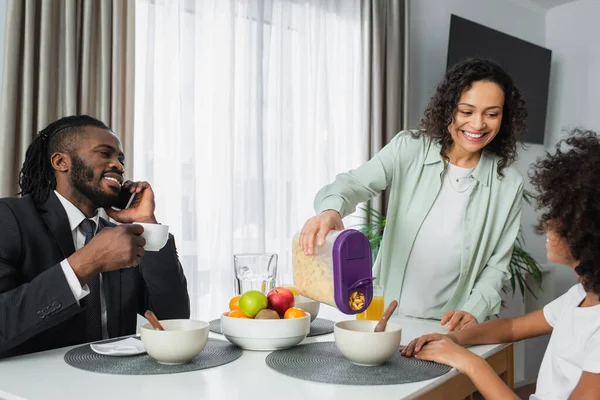 Веселая африканская американка, наливающая кукурузные хлопья в миску дочери рядом с мужем в костюме во время завтрака — стоковое фото