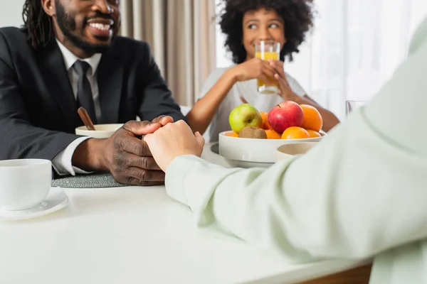 Happy african american man in suit holding hands with wife near daughter during breakfast — Stock Photo