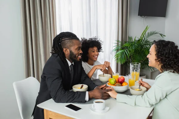 Hombre afroamericano feliz en traje de la mano con la esposa rizada cerca de hija preadolescente durante el desayuno - foto de stock