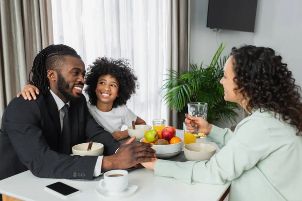 Cheerful african american man in suit holding hands with curly wife near preteen daughter during breakfast — Stock Photo
