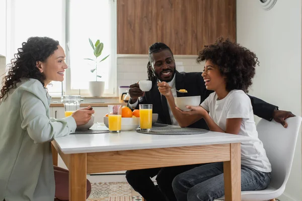 Joyeux parents afro-américains souriants et regardant la fille préadolescente prendre le petit déjeuner dans la cuisine — Photo de stock