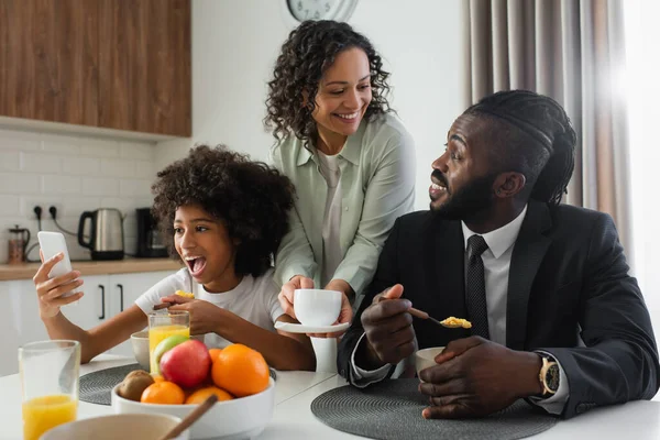 Allegra donna afro-americana che tiene in mano una tazza di caffè vicino al marito in giacca e cravatta mentre la figlia preadolescente utilizza lo smartphone — Foto stock