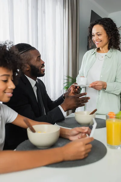 Glücklich afrikanisch-amerikanische Frau hält Tasse Kaffee in der Nähe Ehemann im Anzug und lockige Tochter mit Smartphone — Stockfoto