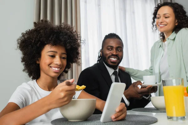 Chica afroamericana feliz usando el teléfono inteligente durante el desayuno con los padres - foto de stock