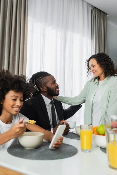 Feliz africana americana mujer abrazando marido cerca hija usando smartphone durante el desayuno - foto de stock