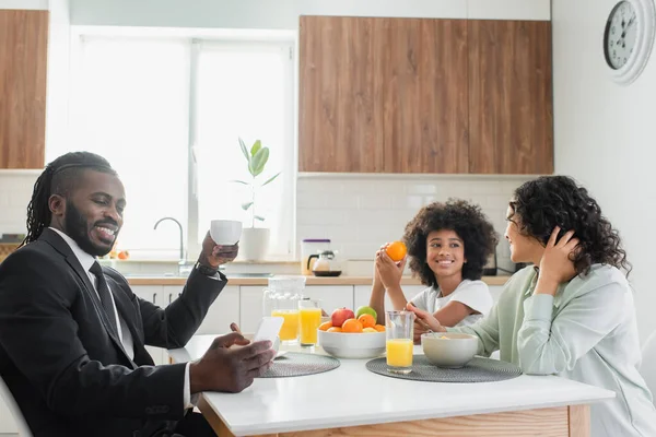 Щасливий афроамериканський чоловік у костюмі, використовуючи смартфон під час сніданку з сім'єю — стокове фото