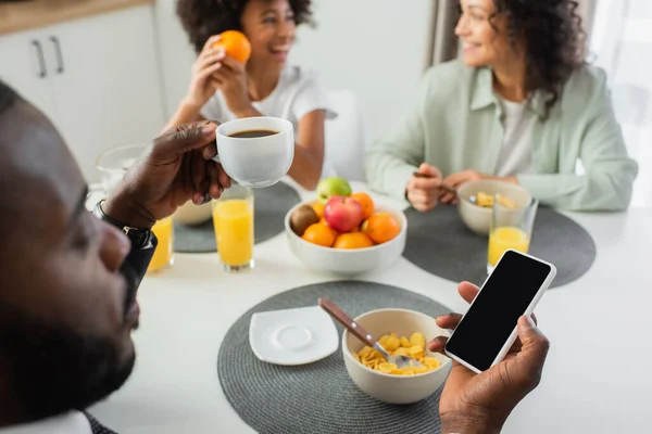 Afrikanisch-amerikanischer Mann benutzt Smartphone mit leerem Bildschirm beim Frühstück mit Familie auf verschwommenem Hintergrund — Stockfoto