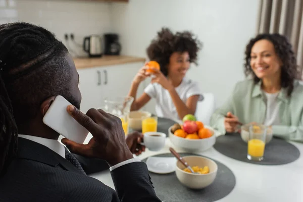 Africano americano empresário falando no smartphone durante o café da manhã com a família em fundo embaçado — Fotografia de Stock
