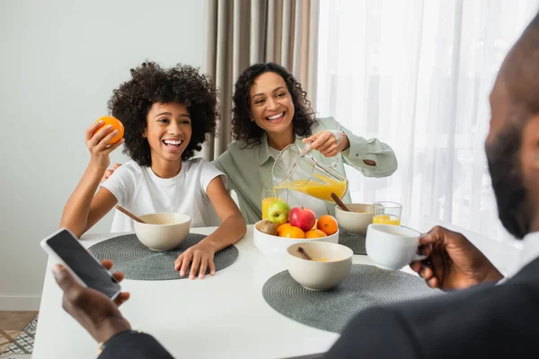 Happy african american woman looking at husband with smartphone near kid during breakfast — Stock Photo
